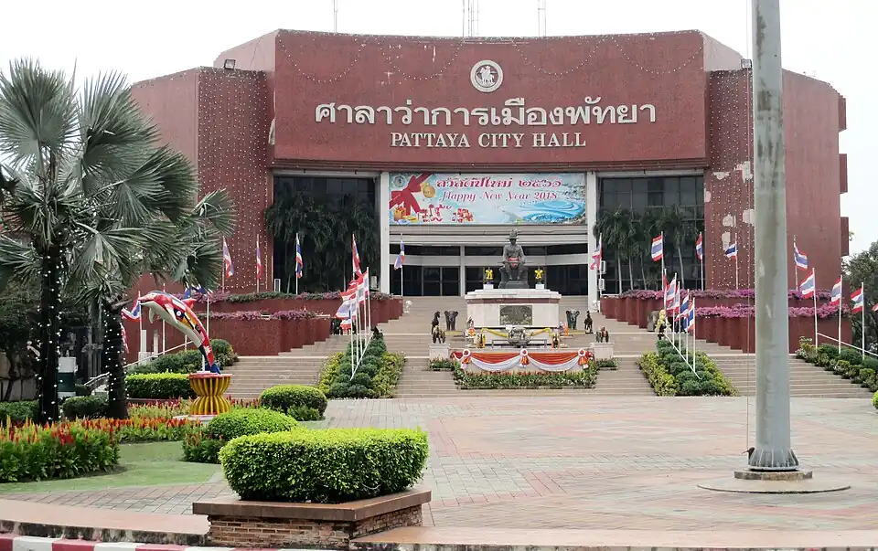 The front of Pattaya City Hall, a red building with its name in Thai and English, flower beds and a flagpole before it