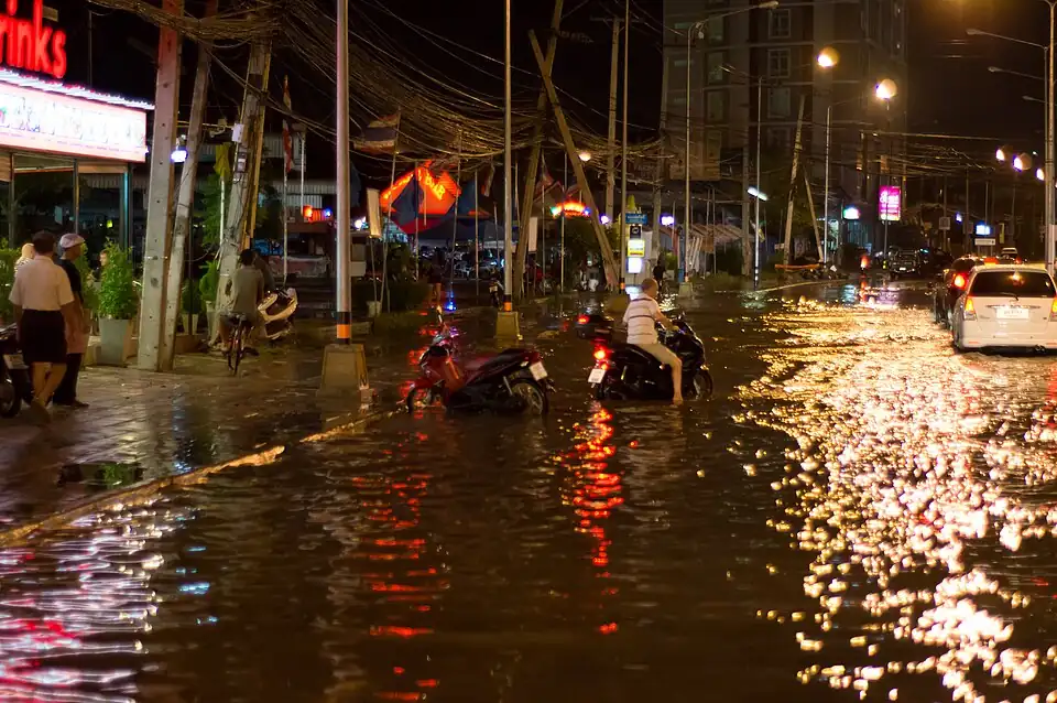 A street in Pattaya at night under ankle-deep floodwater, lights reflecting on the water, motorbikes wading through