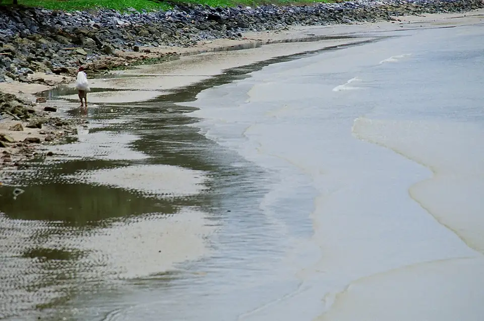 Wet sand and shallow water along Sattahip Beach at low tide, rocks at the top
