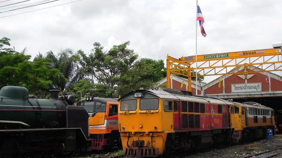 Yellow and orange diesel locomotives and an old steam engine at a Bangkok rail yard under a Thai flag